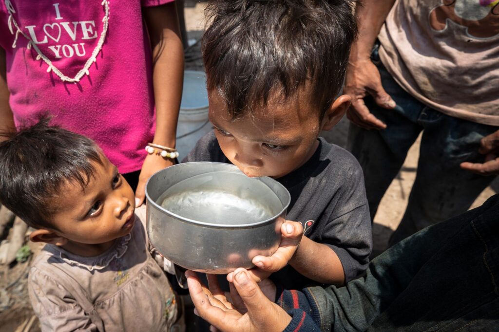 Un enfant boit de l’eau dans un bol en métal.